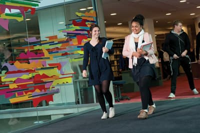 Auf dem Weg zum Studienabschluss Zwei Studenten laufen einen Gang mit bunter Glasfassade entlang.